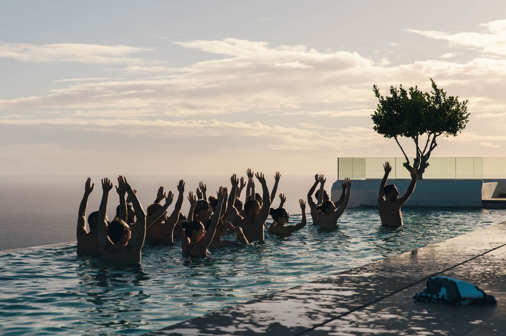 A group gathered in the infinity pool at sunset, hands raised together.