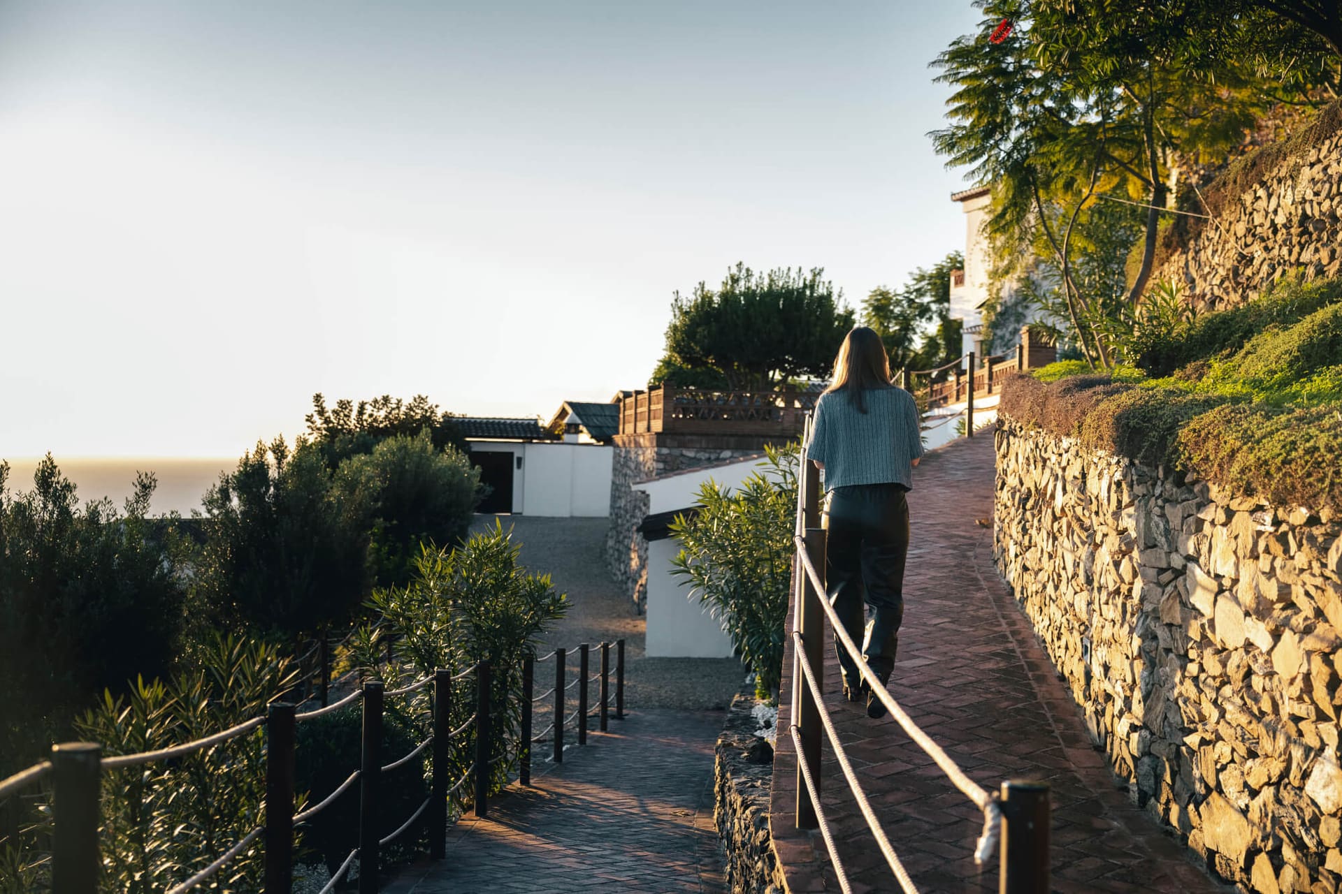A guest walking up the stone-walled path between the villas at sunset, the sea beyond.
